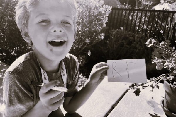 A black and white photo of a little boy laughing holding a marker and a paper with a capital R and lower case r on it.