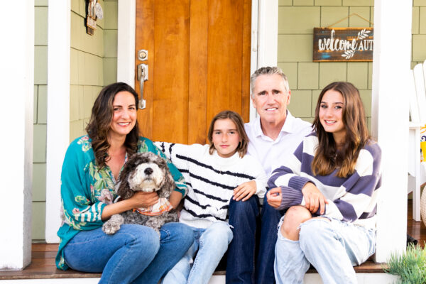 A family with a mom, dad, two daughters and a dog sitting on the porch of a green and white house.