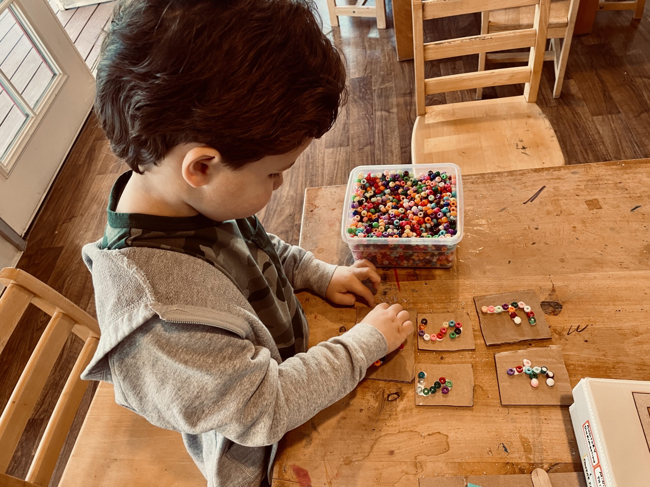 A little boy in a grey hoodie gluing beads on cardboard to form letters.
